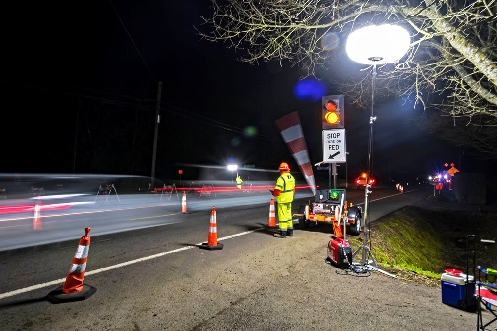 Road construction worker in high-visibility clothing directing traffic at a nighttime work zone with traffic cones, warning signs, and bright work lights.